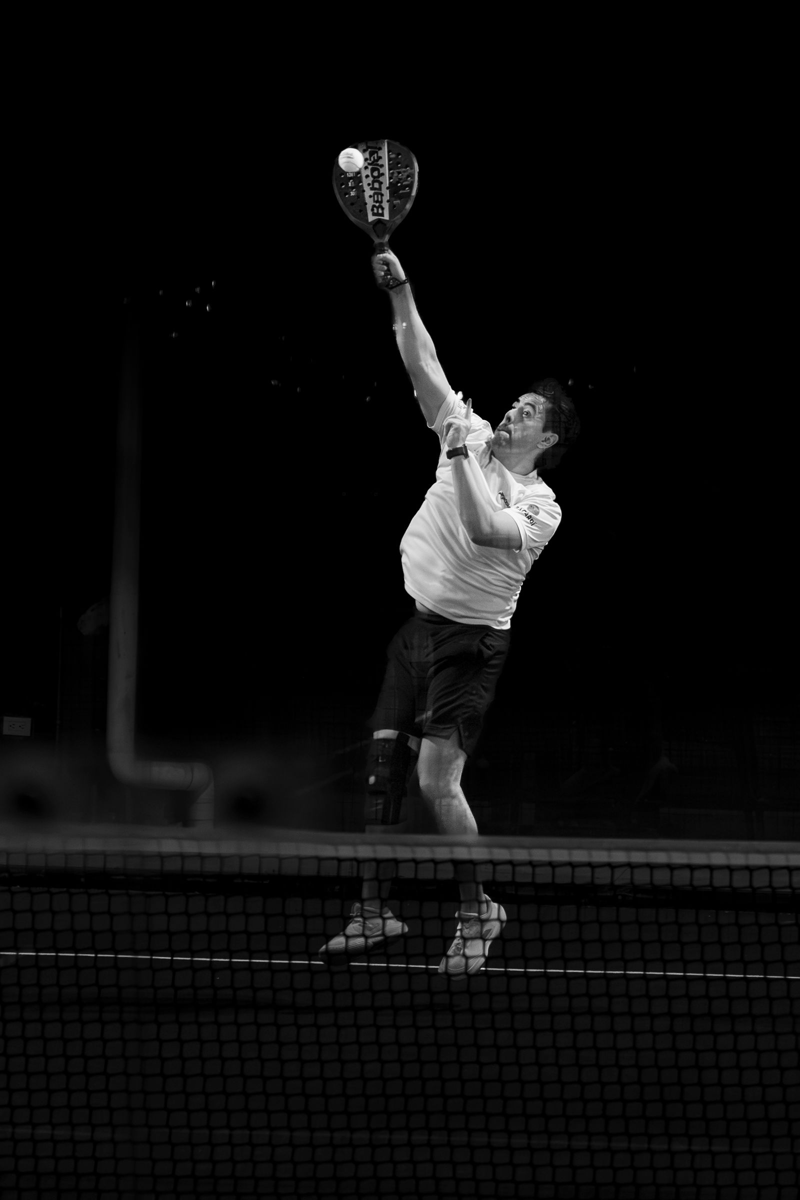 ผู้เล่น padel กระโดดตบเหนือเน็ตในคอร์ทกลางคืน ภาพขาวดำคอนทราสต์สูง