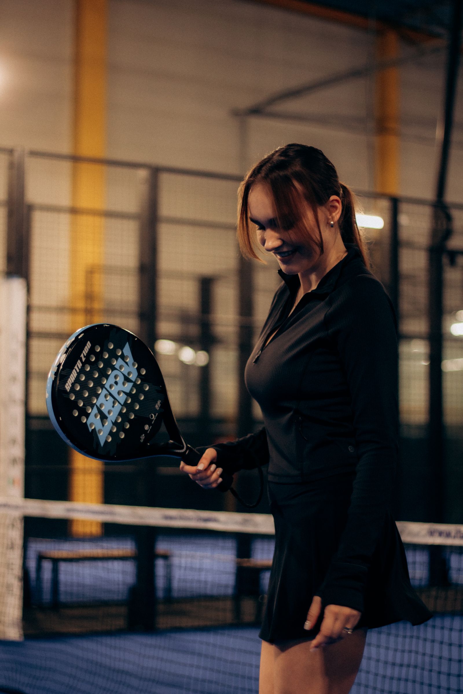 ผู้เล่นหญิงยิ้มมองแร็กเก็ต padel ในคอร์ทแสงอุ่นโทนมืด บรรยากาศผ่อนคลายหลังเล่น